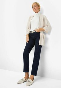Kimono giacca beige chiaro su un dolcevita bianco, abbinato a jeans blu scuro e mocassini crema con accenti metallici.