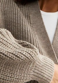 Pletený cardigan v světle hnědé barvě, s texturovaným vzorem, výstřihem do V a žebrovanými manžetami, zdůrazňujícími jemnost látky.