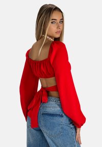 Top rosso corto con maniche a sbuffo e schiena scoperta, legato in vita. Presenta una texture liscia. Abbinato a jeans a vita alta.