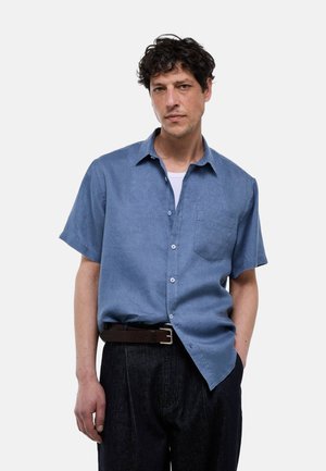 Homme aux cheveux bouclés et foncés portant une chemise bleue à manches courtes boutonnée sur un sous-vêtement blanc et un pantalon noir avec une ceinture marron.