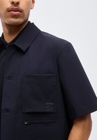 Homme portant une chemise à manches courtes bleu marine foncé avec une grande poche sur la poitrine et une petite poche zippée en dessous.