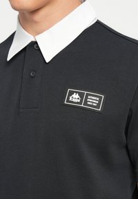 Tricou polo negru cu guler alb, având un patch cu logo-ul Kappa pe piept. Materialul este neted și texturat, cu detaliu cu nasturi.