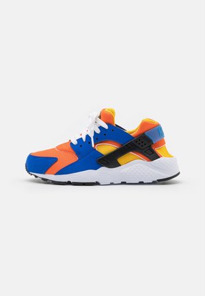 Lavtop atletiksneaker med orange, blå, gule og sorte paneler, hvide snørebånd og en hvid tekstureret sål på en ensfarvet baggrund.