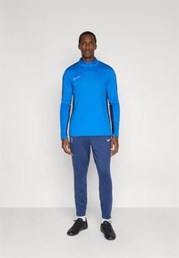 Modrý športový top s dlhými rukávmi a zipsovým golierom, kombinovaný s tmavomodrými nohavicami s logom. Čierne športové topánky, biele ponožky. Jednoduchý dizajn.