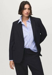Černý pruhovaný blazer s přizpůsobeným střihem, doplněný světle modrou pruhovanou košilí s knoflíky. Obsahuje jednu kapsu a klopy.