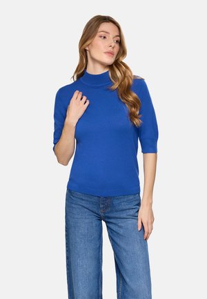 Mujer con cabello largo y ondulado que lleva un suéter azul real de cuello alto y manga corta, y vaqueros azules, de pie frente a un fondo liso.