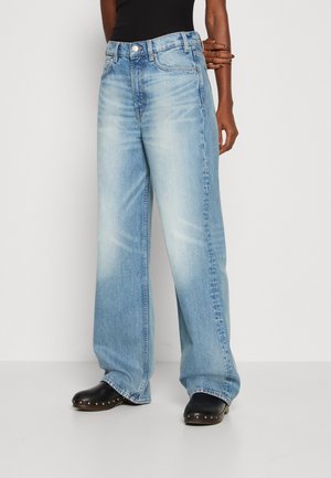Personne portant un jean bleu clair à jambes larges et des sabots noirs avec des clous dorés, debout devant un fond blanc uni.