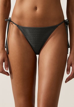 Gros plan d’une personne portant un bas de bikini noir texturé avec des liens latéraux, montrant sa taille et le haut des cuisses sur un fond clair.