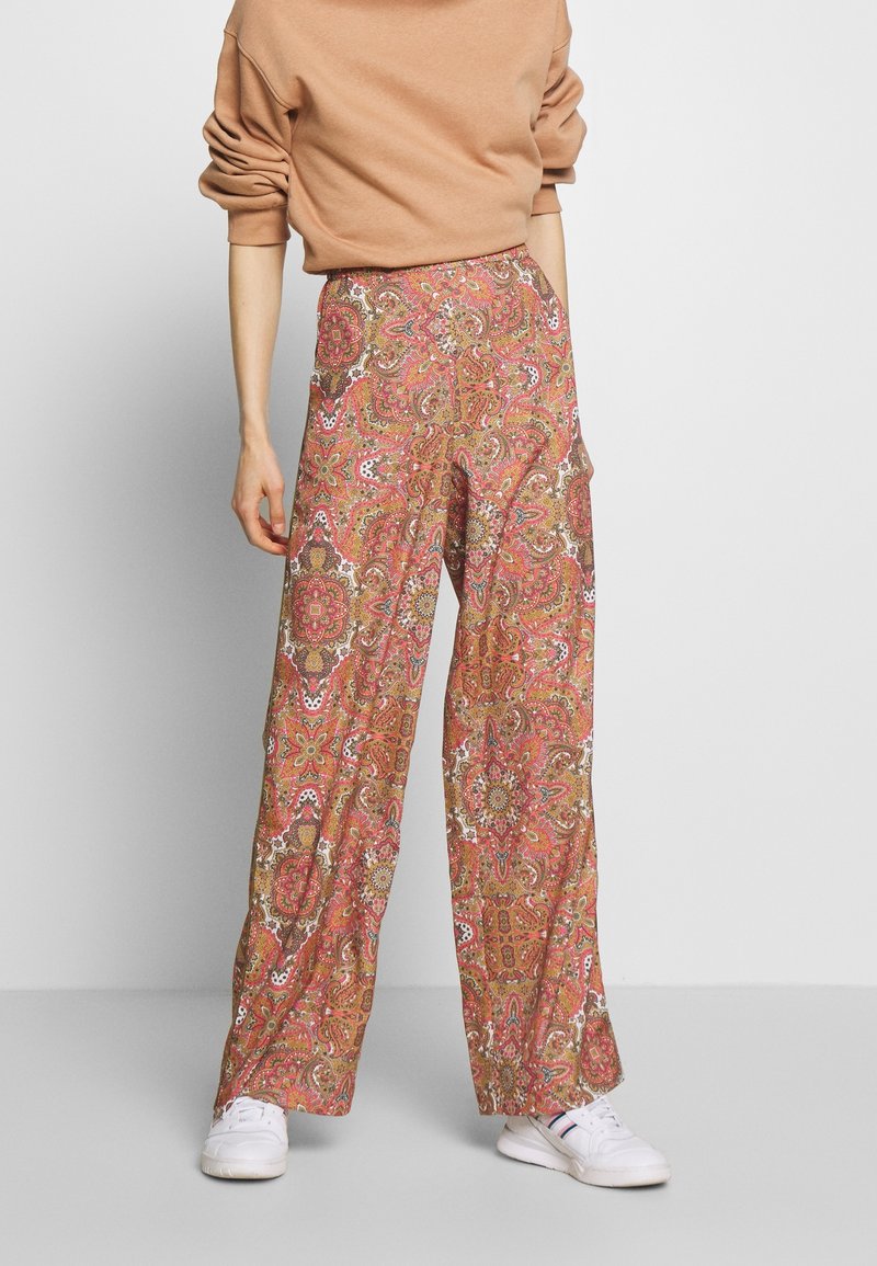 Personne portant un pantalon à jambes larges avec un imprimé floral cachemire rouge, marron et blanc, un haut à manches longues beige et des baskets blanches, debout sur un sol gris.