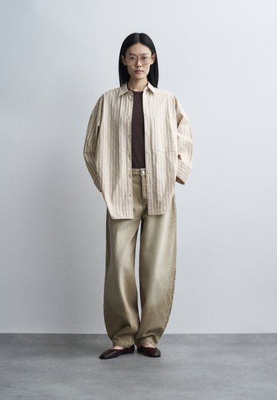 Jeune femme portant des lunettes rondes, une chemise beige rayée ample sur un haut sombre, un pantalon beige large et des chaussures marron, debout contre un mur gris uni.