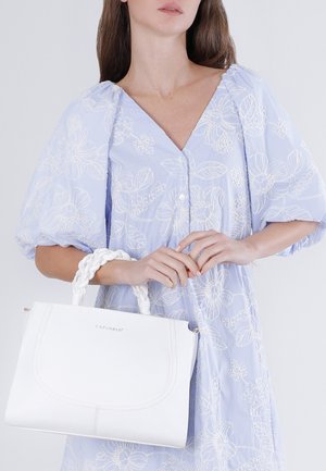 Vaaleansiniseen kukkakuosiseen mekkoon pukeutunut nainen, jonka hihassa on pussimaiset hihat, pitää valkoista nahkaista käsilaukkua, jossa on punotut kahvat.