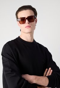 Négyzet alakú napszemüveg sötétbarna lencsékkel, vastag barna kerettel és ezüst színű kiegészítőkkel. A modellen fekete, kerek nyakú pulóver van.