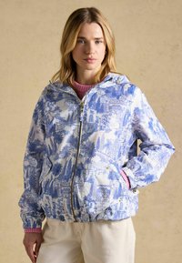 Naine, kes kannab valget lukuga jopet sinise linnavaate mustriga ja heleroosa kampsunit all, seisab ühe käega taskus.