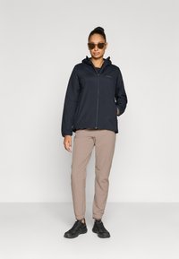 Casaco leve azul-marinho com capuz, fecho de correr frontal, punhos elásticos; combinado com calças bege e sapatos pretos, em pé sobre um fundo neutro.