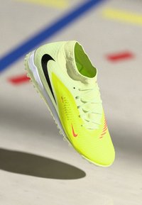 Zapatilla deportiva en amarillo brillante y blanco con swooshes de Nike en negro y rosa, cordones y suela texturizada sobre un fondo de una cancha interior.