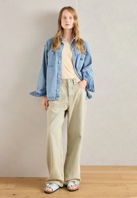 Denim srajca v svetlo modri barvi, bež majica, svetlo zelene hlače širokega kroja in modri natikači z zaponko, stojijo na lesenem tlu.