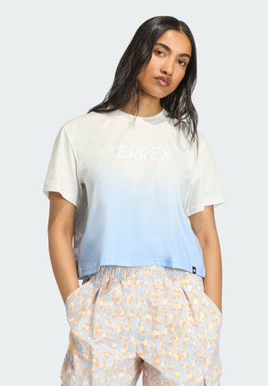 Γυναίκα με μακριά σκοτεινά μαλλιά, που φοράει ένα t-shirt TERREX με ombre από λευκό σε ανοιχτό μπλε και ψηλόμεσα παντελόνια με πορτοκαλί μοτίβο λεοπάρδαλης.