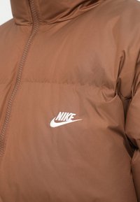 Smeđi prošiveni jakna s prednjim patentnim zatvaračem i bijelim Nike logotipom izvezenim na lijevoj strani grudi.