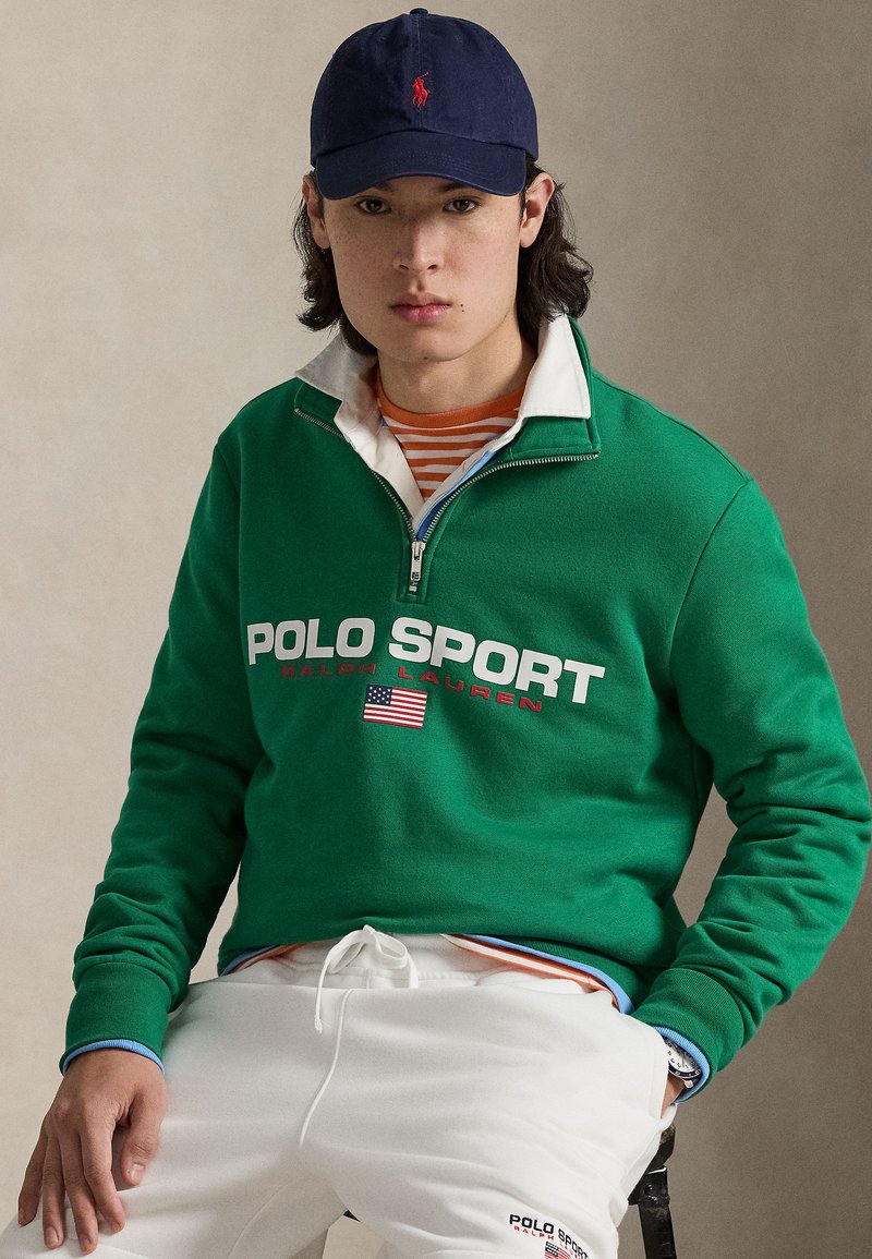 Green zip-up sweatshirt with "POLO SPORT" text and American flag detail, layered over a striped shirt, paired with white pants and a navy cap.