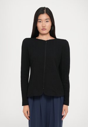 Woman with straight black hair wearing a textured black zip-up jacket and loose navy blue pants against a plain white background.