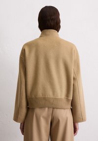 Beige Wolljacke im Cropped-Style mit hohem, geripptem Kragen und weiten Ärmeln. Glatte Textur mit einem taillierten, gerippten Saum. Einfaches Design, ohne Muster.