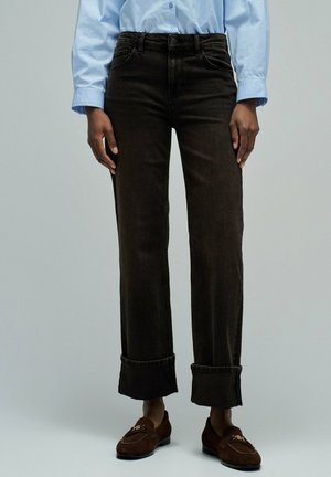 Persona vestida con vaqueros negros con puños, mocasines marrones y una camisa azul clara abotonada, de pie frente a un fondo liso.