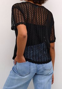Czarny top na szydełku z krótkimi rękawami, z geometrycznym wzorem i zakończeniem z falbanką. Połączony z jasnoniebieskimi dżinsami, prezentując swobodny styl.