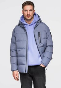 Szara kurtka puchowa z kapturem, z zamykaną na zamek kieszenią na piersi i elastycznymi ściągaczami przy mankietach. Nosi się ją na jasnofioletowej bluzie z kapturem i ciemnych dżinsach.