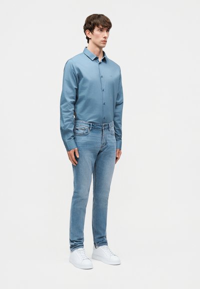 Άνδρας που στέκεται φορώντας μπλε πουκάμισο με κουμπιά, ανοιχτόχρωμο μπλε τζιν και λευκά αθλητικά παπούτσια, μπροστά σε απλό φωτεινό φόντο.