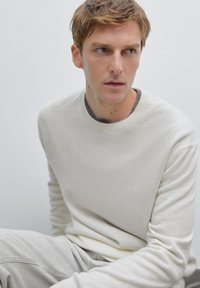 Hellbeige gestrickter Pullover mit Rundhalsausschnitt, gerippten Bündchen und weicher Textur, kombiniert mit hellgrauen Hosen, vor einem neutralen Hintergrund.