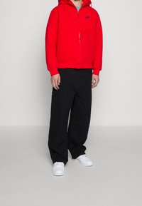 Person som bär en röd Nike-hoodie, svarta vida byxor och vita sneakers, stående mot en enfärgad grå bakgrund.