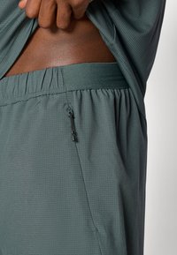 Αθλητικές βερμούδες από υφή σκούρου πράσινου υφάσματος, με ελαστική μέση και μια μικρή τσέπη με λεπτομέρεια φερμουάρ.