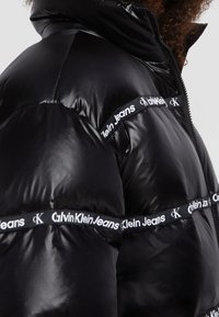 Veste en duvet noire avec une finition brillante, présentant des détails de bande avec le logo "Calvin Klein Jeans" en blanc sur les côtés et un col montant. Fermeture éclair.