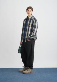 Ilga rankovėms marškiniai su languotu raštu tamsiai mėlyno ir kreminio atspalvio, derinami su juodomis plačiomis kelnėmis ir kreminės spalvos batais. Laikydama žalią rankinę, stovi ant mėlynų grindų.