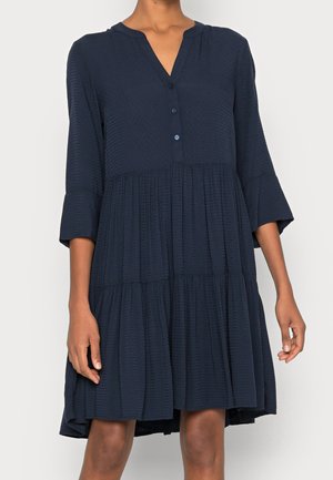 Granatowa sukienka z rękawami do łokcia, dekoltem w serek, guzikami i falbanami ułożonymi warstwowo. Wykonana z fakturowanej tkaniny, o długości do kolan.