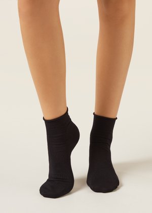 Nohy obuté v černých kotníkových ponožkách, stojící na špičkách na neutrálním pozadí.