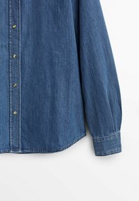 Chemise en denim bleu à manches longues avec boutons-pression métalliques et ourlet arrondi, présentée sur un fond blanc uni.