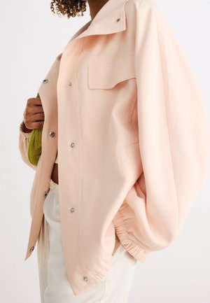 Un mannequin porte une surchemise rose pâle oversize avec des boutons-pression et une poche poitrine, tenant un sac vert texturé.