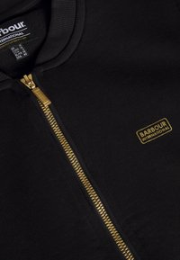 Černá bunda na zip z měkké tkaniny se zlatým zipem, na hrudníku malý zlatý nápis „Barbour International“.