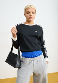 Fekete, hosszú ujjú ing fehér díszítéssel és Adidas logóval, szürke melegítőnadrággal kombinálva, kék elasztikus derékpánttal. Fekete kézitáska látható.