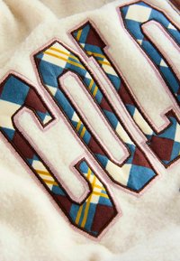 Crème-kleurig fleece stof met grote, meerkleurige "COL" letters met een blauw, bruin, geel ruitpatroon en roze omlijning stiksel.