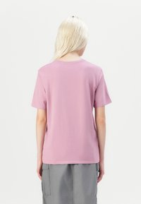 En blond person med rett hår har på seg en kortermet rosa T-skjorte og grå cargo-skjørt, med ryggen mot kameraet mot en hvit bakgrunn.