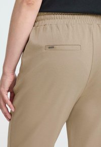 Beige broek met een elastische tailleband, voorzien van een klein, horizontaal achterzakje en een metalen logoaccent op het zakje.
