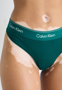 Teal katoenen bikini met een Calvin Klein-logo op de tailleband. Bevat een gladde textuur en een low-rise snit, waarmee variaties in huidtint worden benadrukt.