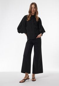 Zwarte oversized shirt met ballonmouwen en een kraag, gecombineerd met zwarte wijde broek. Beide hebben een gladde textuur en een minimalistisch ontwerp.