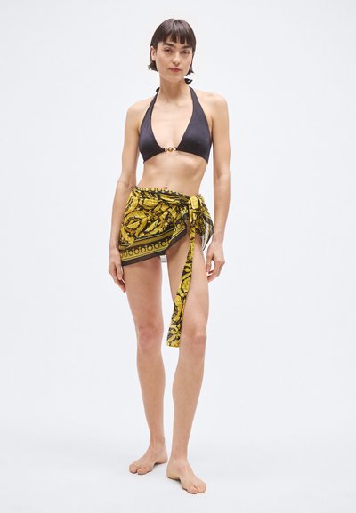 Čierny vrchný diel bikín so zlatými ozdobami, doplnený o žlto-čiernu sarong so vzorom, viazaný v páse. Modelka stojí bosá na neutrálnom pozadí.