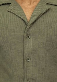 Chemise olive verte à boutons avec un tissu texturé et à motif carré. Elle a un col classique et deux boutons visibles à l'avant.