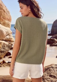 Top texturizado verde oliva con mangas cortas, combinado con pantalones cortos blancos con dobladillo enrollado. El escenario incluye una playa rocosa y un fondo de océano.