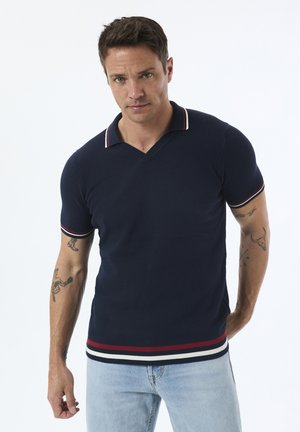 Navyblaues Polohemd mit strukturiertem Gewebe, V-Ausschnitt und kontrastierendem rot-weißen Besatz am Kragen und den Ärmeln, kombiniert mit hellblauen Jeans.