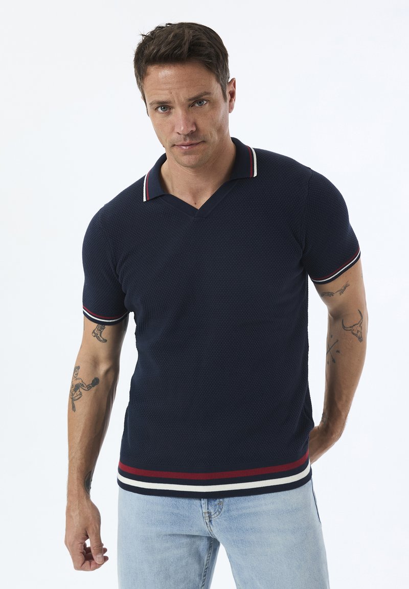 Navyblaues Polohemd mit strukturiertem Gewebe, V-Ausschnitt und kontrastierendem rot-weißen Besatz am Kragen und den Ärmeln, kombiniert mit hellblauen Jeans.
